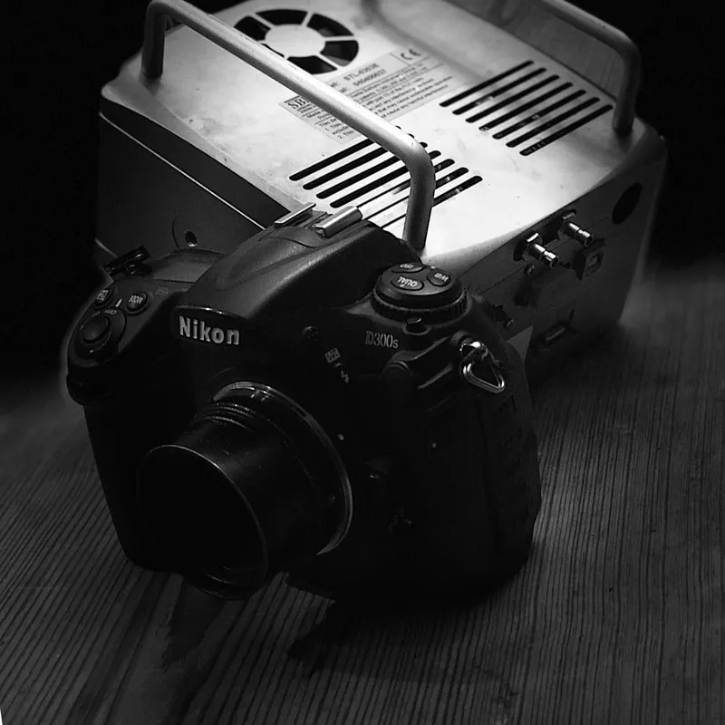 A Nikon D300s DSLR camera photographed alongside a portable power supply, in black and white